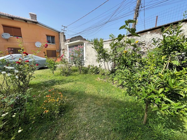 A residential garden with a variety of plants and flowers, a car parked in the driveway, and a building with a stone wall and a metal fence.