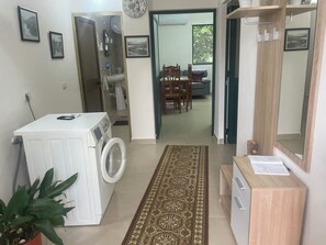 A laundry room with a washing machine, a small potted plant, and a patterned rug leading to a dining area with a table and chairs.