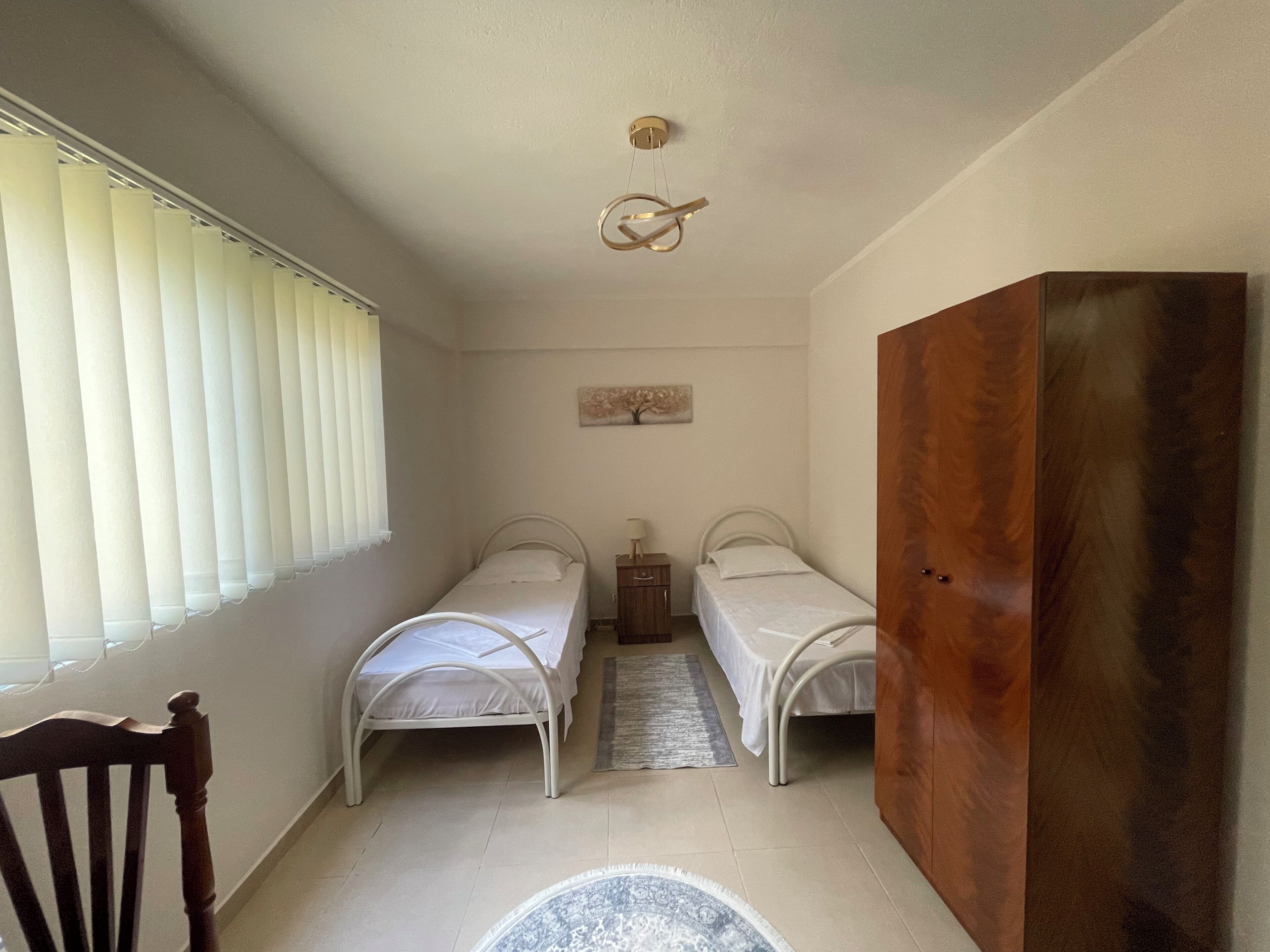A bedroom with two single beds, a wooden wardrobe, a small table, and a ceiling light fixture.