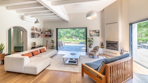 Ein modernes Wohnzimmer mit einer großen Couch, einem Couchtisch, einer Holzbank und einem Kamin. Durch eine Glastür ist ein Bücherregal und ein Essbereich zu sehen.
