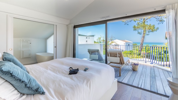 Et soveværelse med en stor seng, en skydedør til en balkon med havudsigt og et træ.