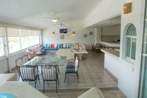 A spacious living area with a glass-top dining table, comfortable seating, a ceiling fan, and a kitchen area in the background. - Apt. with Jacuzzi Ixt. Zihuatanejo 3 BR (Ixtapa)