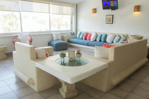 A living room with a sofa, armchairs, a glass-top coffee table, and a TV mounted on the wall. - Apt. with Jacuzzi Ixt. Zihuatanejo 3 BR (Ixtapa)