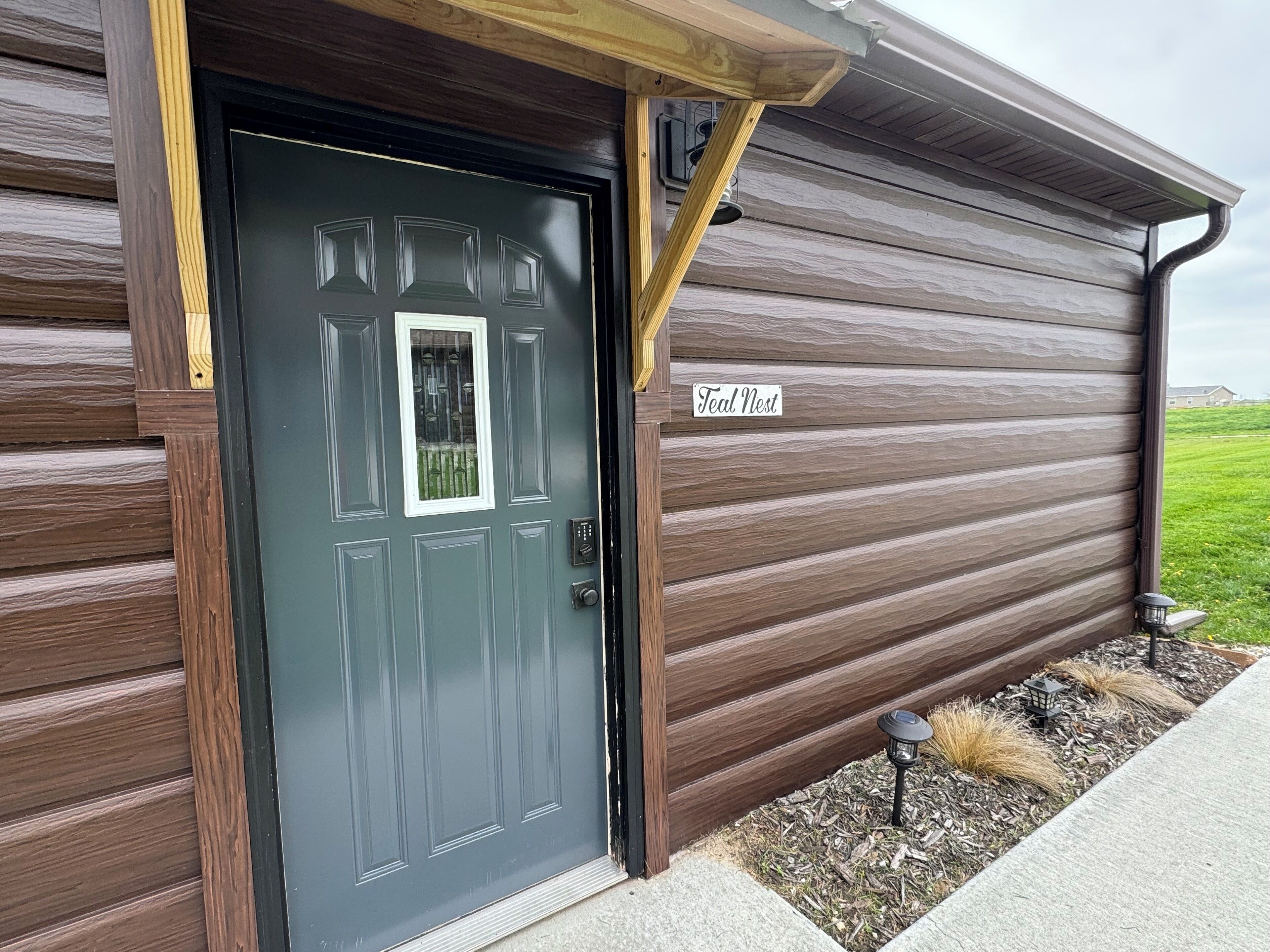 A wooden exterior with a dark grey door, a nameplate reading "Teal Nest," and a small garden area with mulch and plants.