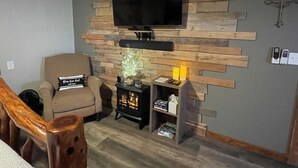 Ein Wohnzimmer mit einem an einer Holzwand montierten Flachbildfernseher, einem beigen Sessel, einem kleinen Tisch mit einer Zierpflanze und einem Regal mit Büchern und Kerzen.