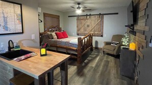 Ein gemütliches Schlafzimmer mit einem hölzernen Bett, einem Schreibtisch und einem Stuhl. Eine Deckenventilator, eine kleine Weihnachtstanne und eine Karte sind an der Wand zu sehen.