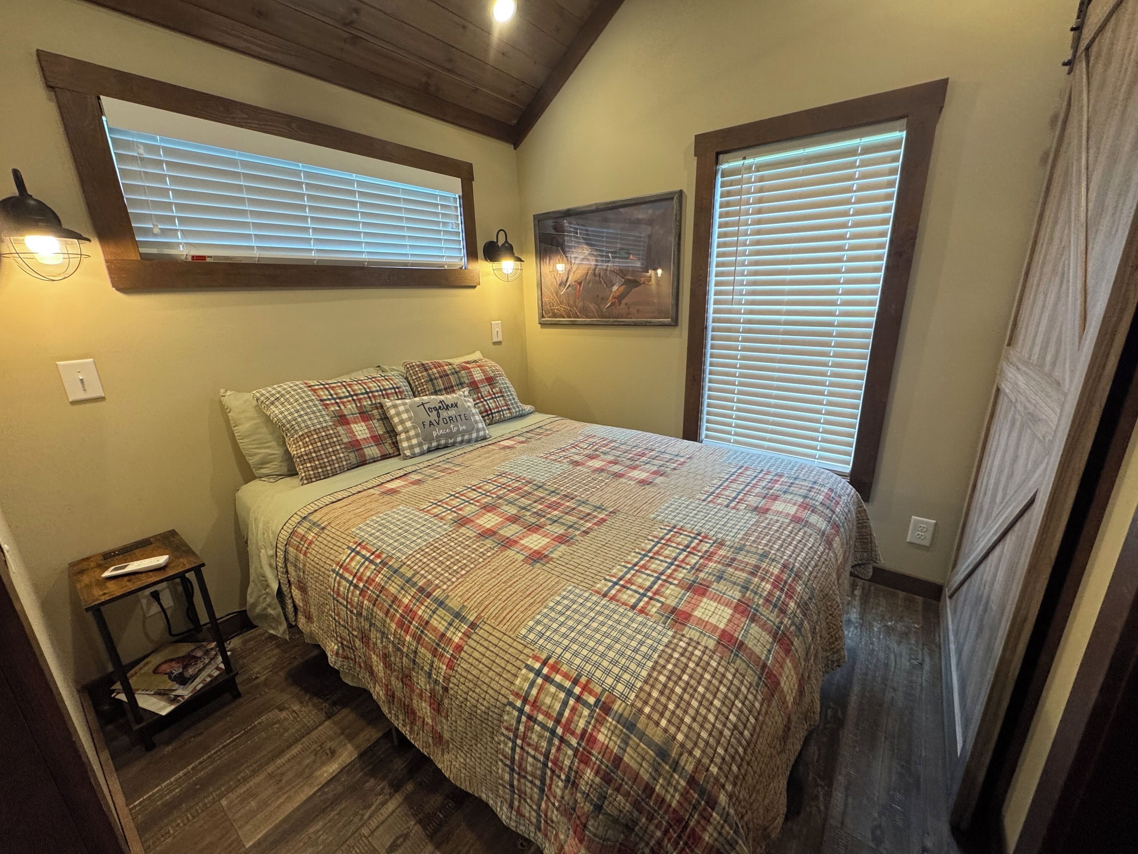 Un dormitorio con cubrecama a cuadros, dos lámparas de noche, un cuadro en la pared y una ventana con persianas.