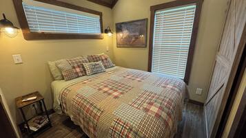 Un dormitorio con cubrecama a cuadros, dos lámparas de noche, un cuadro en la pared y una ventana con persianas.