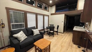 Una sala de estar acogedora con un sofá, una mesa de centro de madera y una mesa de comedor con sillas. Varias ventanas con persianas, una zona de cocina con fregadero y armarios, y un balcón con barandilla.