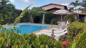 A swimming pool with lounge chairs and an umbrella, surrounded by lush greenery and a building with a red roof. - Cumbuco - Beach house with pool and deck Ceará (Praia de Cumbuco Caucaia)
