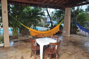 A covered patio with a table and chairs, a hammock, and a pool in the background. - Cumbuco - Beach house with pool and deck Ceará (Praia de Cumbuco Caucaia)
