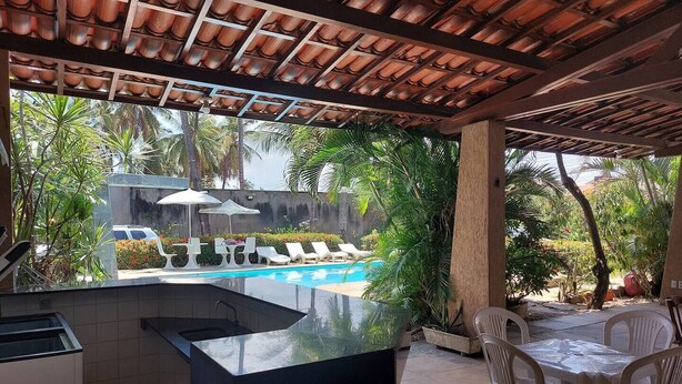 A poolside area with a kitchen counter, outdoor seating, and a view of a pool and waterfall. - Cumbuco - Beach house with pool and deck Ceará (Praia de Cumbuco Caucaia)