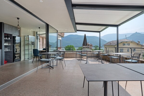 Un espace de restauration extérieur moderne, avec des tables et des chaises, un bar doté d’étagères et une vue sur les montagnes.