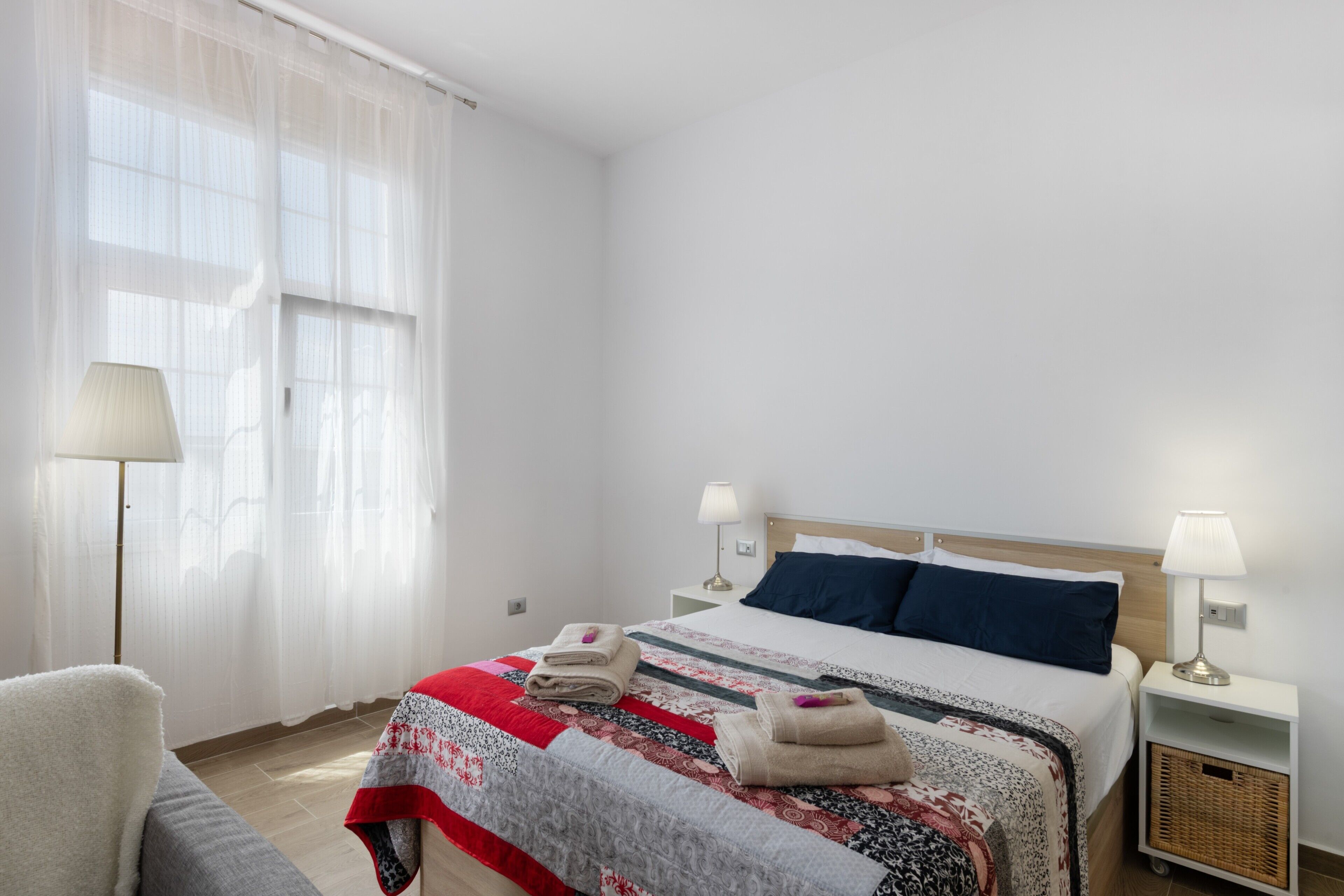 A hotel room with a bed, two bedside lamps, a chair, a lamp, and a window with sheer curtains.