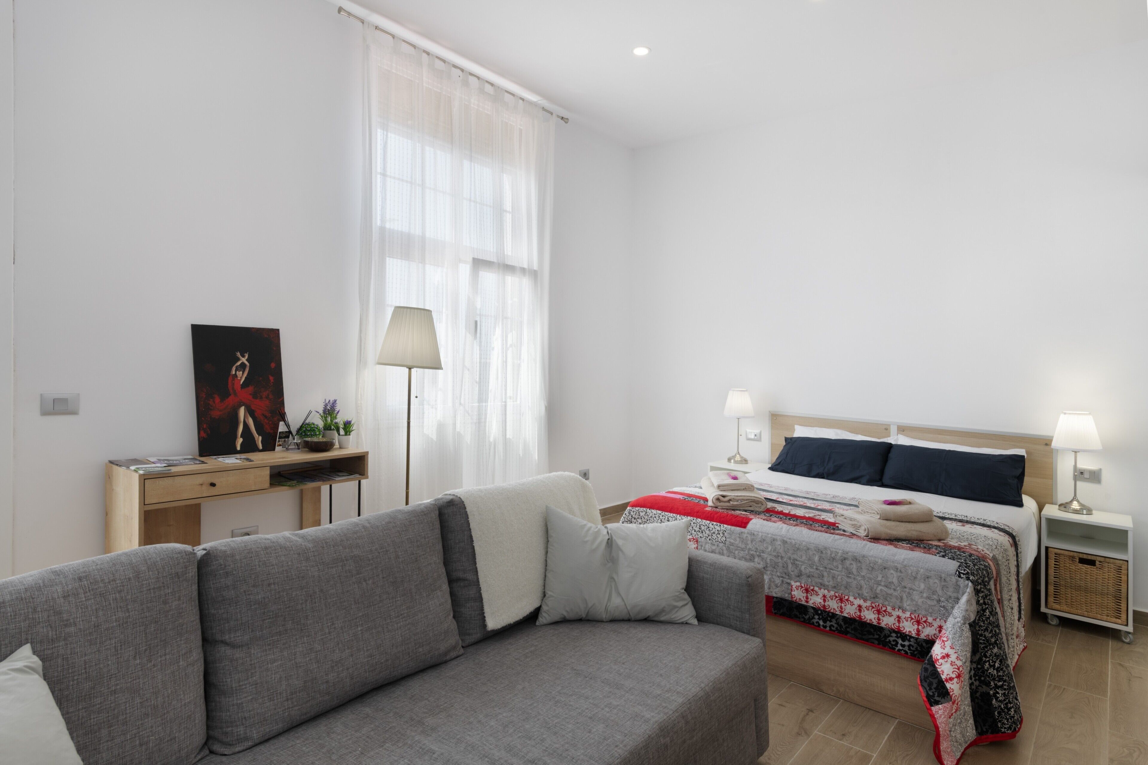 A modern bedroom with a grey sofa, a bed with a red and black patterned bedspread, a bedside table with a lamp, and a desk with a lamp and a framed picture.