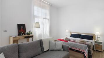 A modern bedroom with a grey sofa, a bed with a red and black patterned bedspread, a bedside table with a lamp, and a desk with a lamp and a framed picture.