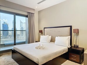 Hotelkamer met een groot bed, nachtkastje, lamp en uitzicht op een stadslandschap door een groot raam.