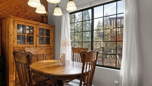 A wooden dining table with six chairs, a cabinet with glass doors, and a large window with a view of trees and a wooden deck. - Little Bear Lodge (Big Bear Lake)