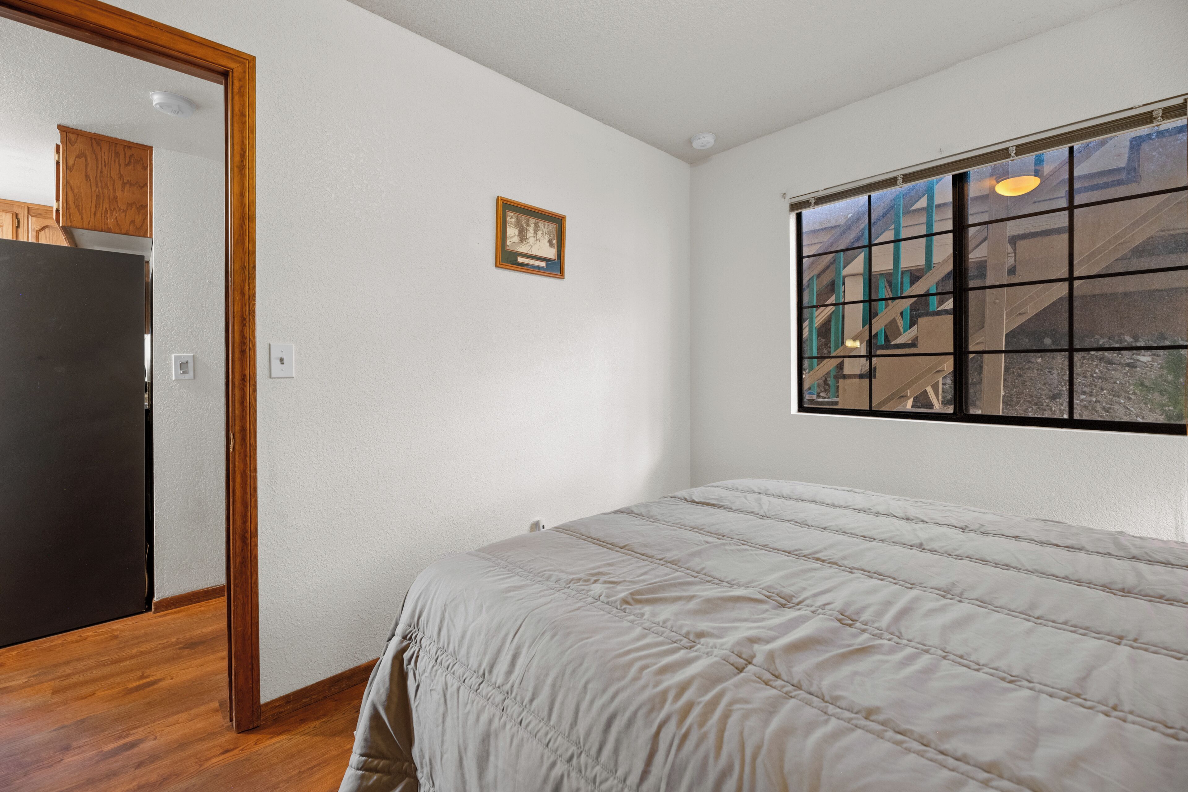 Una camera da letto con un letto, pavimento in legno, pareti bianche, una finestra vista scale e una porta che dà su un'altra stanza.