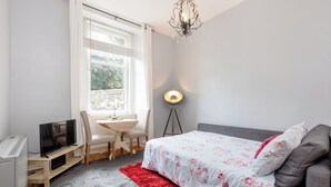 Ein Schlafzimmer mit einem Bett, einem Schreibtisch mit Computer, einem Fernseher, einer Lampe und einem Fenster mit Vorhängen.