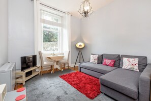 Ein kompakter Wohnbereich mit grauem Sofa, rotem Teppich, einem kleinen Esstisch und einem Fernseher.
