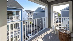 A balcony with a white chair and a wicker table, overlooking a parking area and neighboring houses.