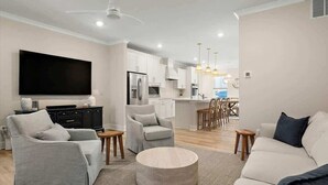 A modern living room with a sofa, armchair, and ottoman, a flat-screen TV, and a kitchen in the background.