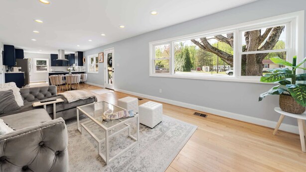 A spacious living area with a large window, a grey sofa, a coffee table, and a kitchen in the background. - Coastal Modern Retreat | Large Group Getaway on 2 Acres (Arnold)