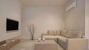 Ein modernes Wohnzimmer mit einem beigefarbenen Ecksofa, einem Flachbildfernseher an der Wand, einem kleinen Beistelltisch und einer dekorativen Vase mit Zweigen.