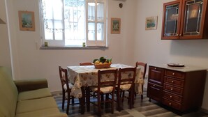 Un coin repas avec une table dressée pour quatre personnes, entourée de chaises en bois recouvertes d’un tissu à motifs floraux. Un meuble en bois avec des tiroirs se trouve à droite.