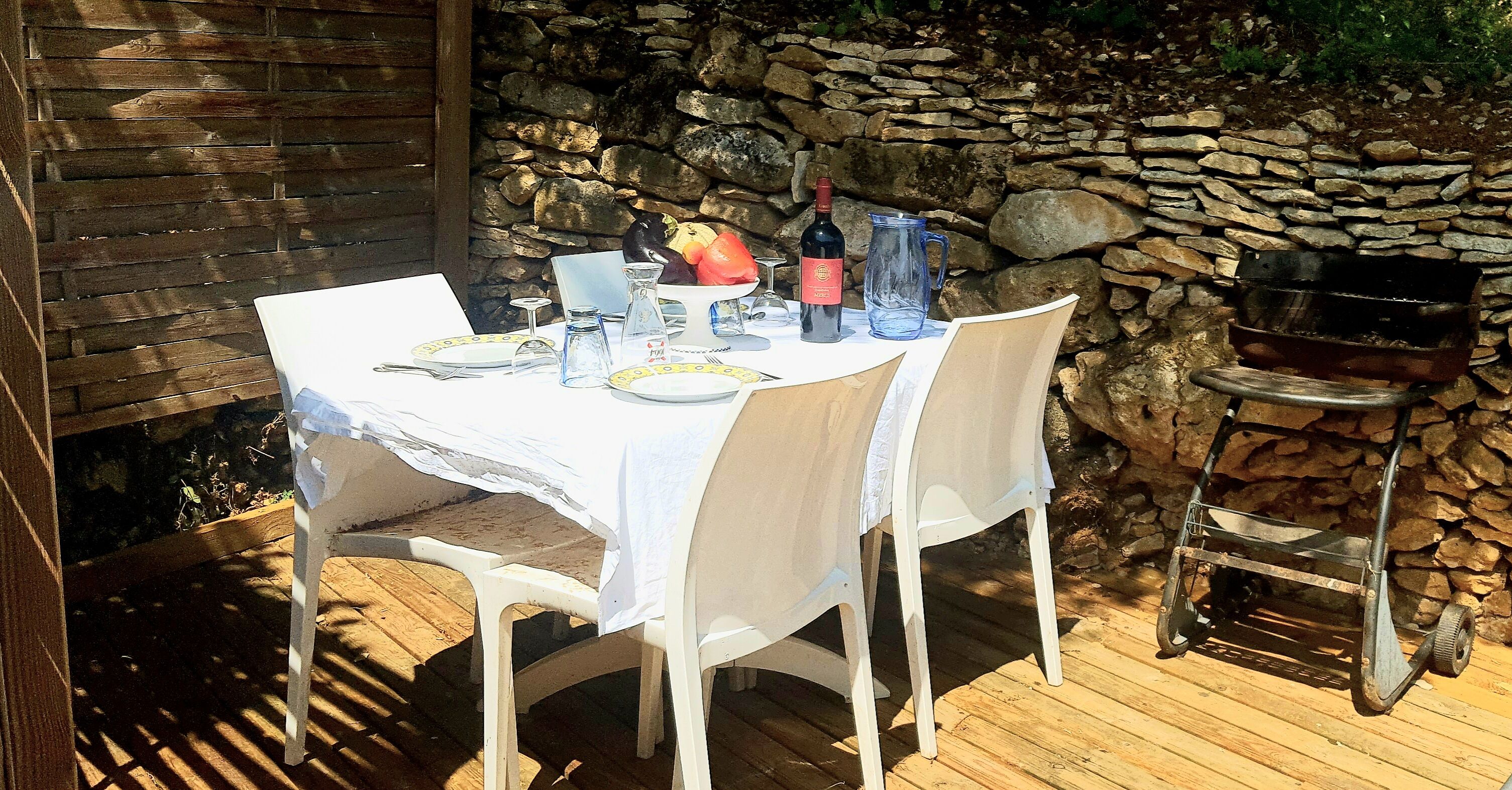 Uma área de refeições ao ar livre com um muro de pedra ao fundo, uma churrasqueira e uma mesa posta com pratos, copos e uma garrafa.
