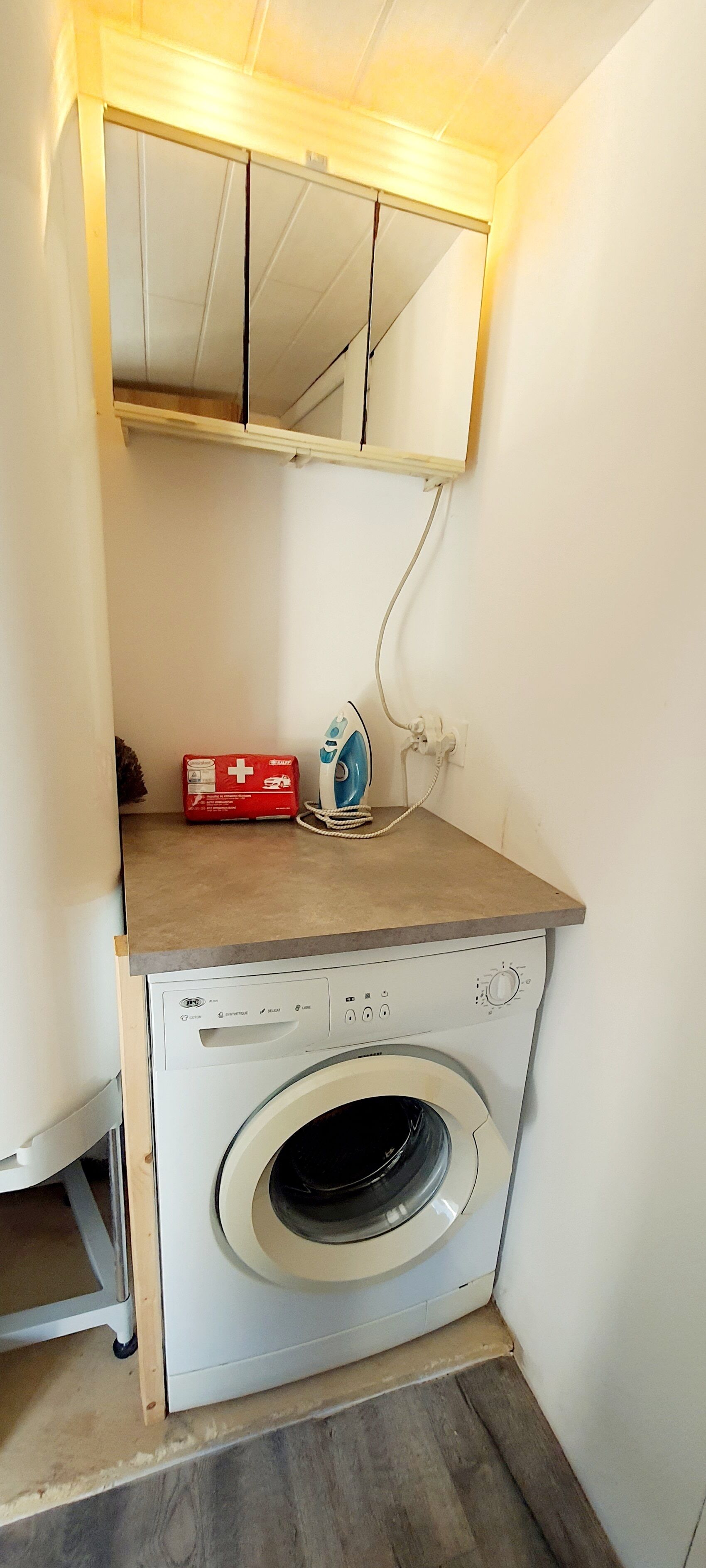 Un espace réduit dédié à la buanderie, comprenant un lave-linge, une trousse de premiers secours rouge et un fer à repasser bleu, le tout posé sur un plan de travail en bois.