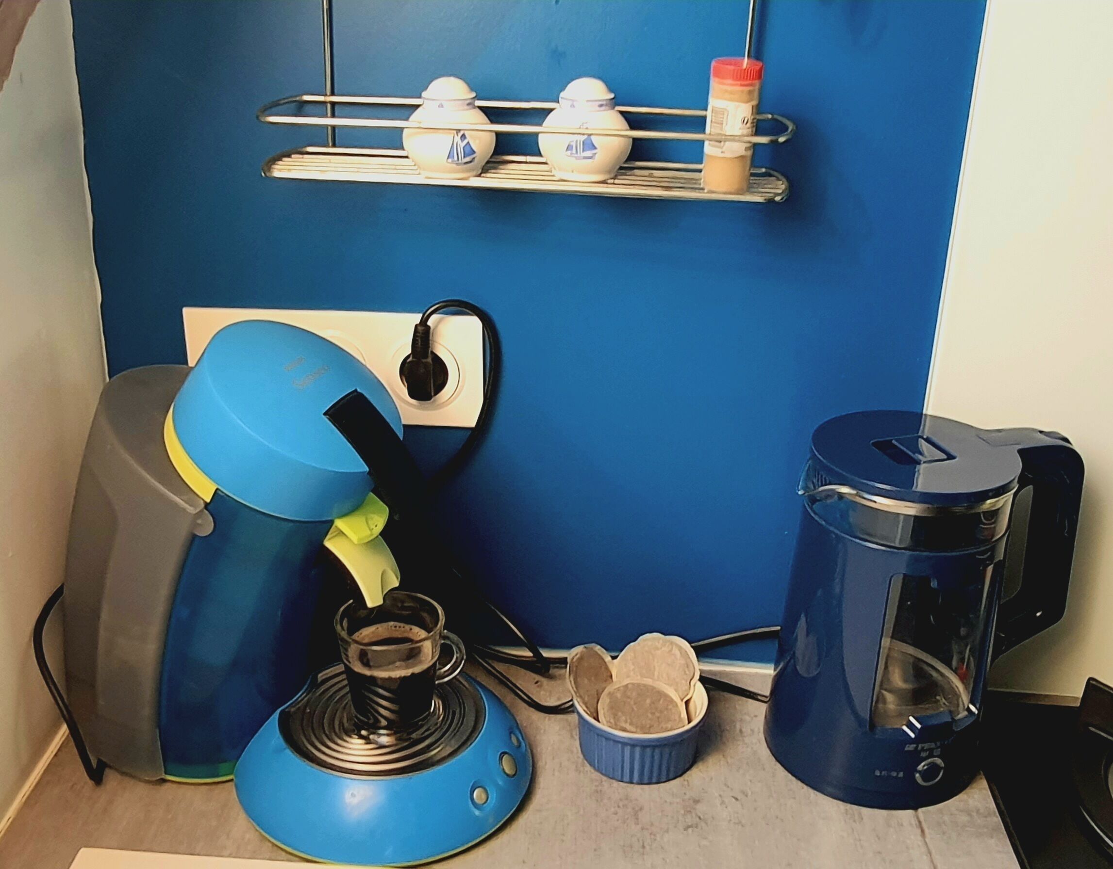 A kitchen with a coffee maker, a kettle, and a toaster. There is a shelf above the counter with cups and a condiment bottle.