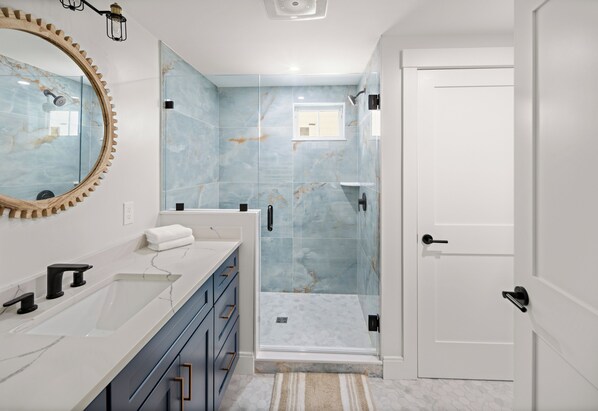 A bathroom with a marble shower wall, a large mirror, a sink with a towel, and a door.
