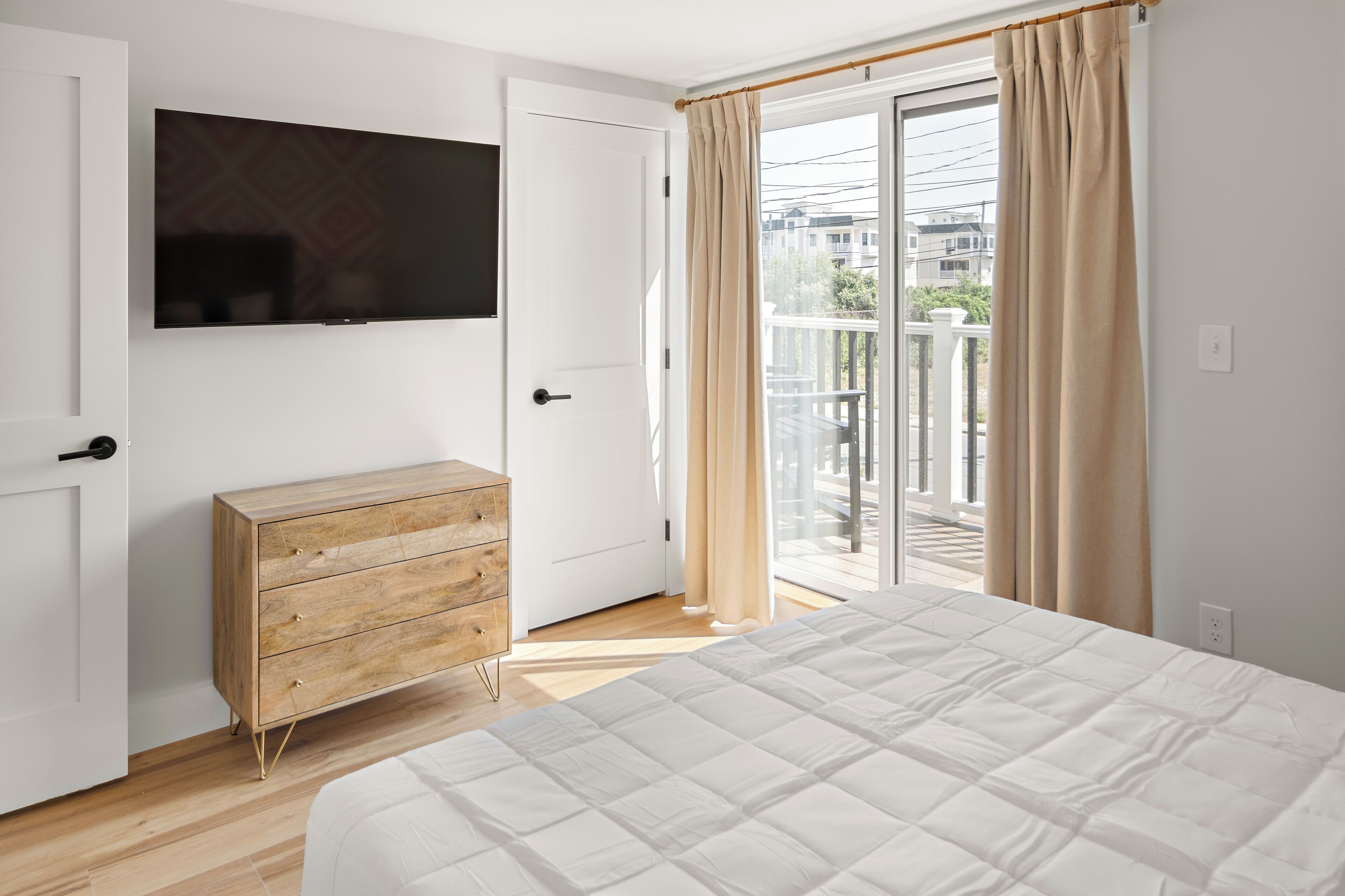 A bedroom with a bed, a wooden dresser, a television on the wall, and a sliding glass door to a balcony.