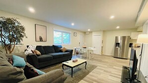 Una sala moderna con un sofá, una mesa de centro y un mueble para televisión. Hay una zona de comedor con una mesa y sillas, y una cocina con un refrigerador.