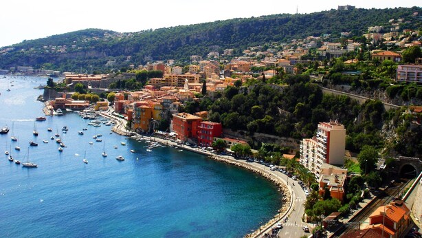A coastal town with buildings along the waterfront, a marina with boats, and a hilly backdrop.