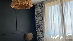 A bedroom with a dark wall, a large bed with grey bedding, a woven light fixture, and sheer curtains.