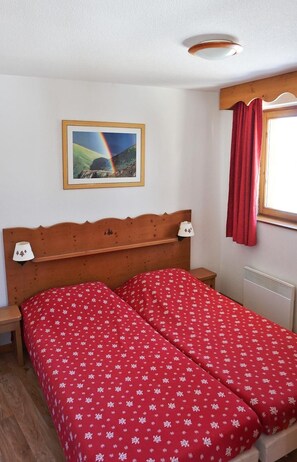 A double bed with red floral bedding, a wooden headboard, a bedside lamp, a framed picture of a rainbow, and a window with red curtains.