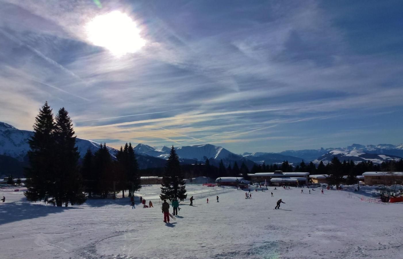 Ein verschneiter Skigebiet mit blauem Himmel, Bergen im Hintergrund und mehreren Gebäuden, vermutlich für Unterkünfte oder Dienstleistungen.