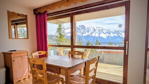 A wooden dining table with chairs, a wooden cabinet, and a large window with a view of snow-capped mountains. - 70m from ski slopes, fitness, swimming pool, sauna, terrace, balcony, parking, tv, 25m², Chamrousse (Chamrousse)