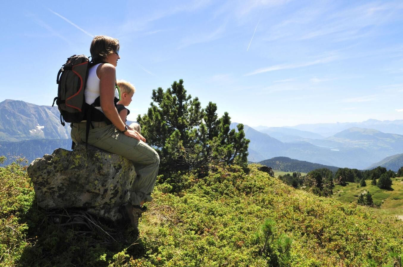 A person sitting on a rock with a backpack, overlooking a mountainous landscape with trees and a clear sky.