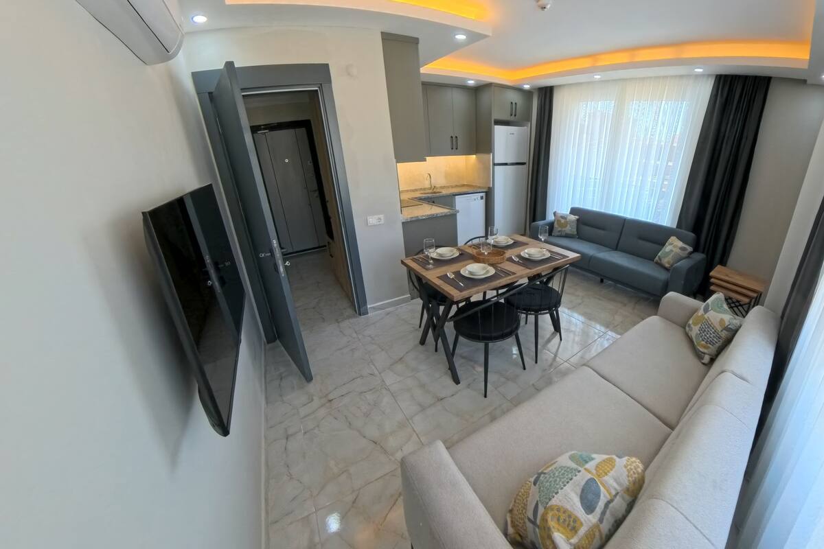 A modern living room with a sofa, a dining table set with plates and glasses, a kitchen area with cabinets and a refrigerator, and a TV mounted on the wall.
