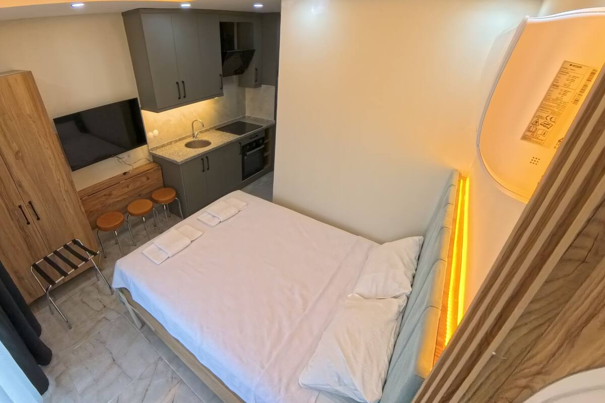 A compact hotel room with a bed, a small kitchenette, a wooden wardrobe, and a seating area with stools.