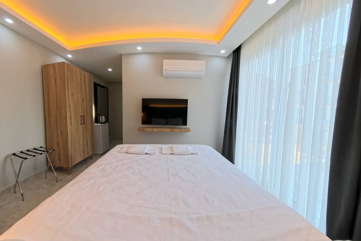 A modern hotel room with a large bed, a wooden wardrobe, a TV mounted on the wall, and a window with sheer curtains.