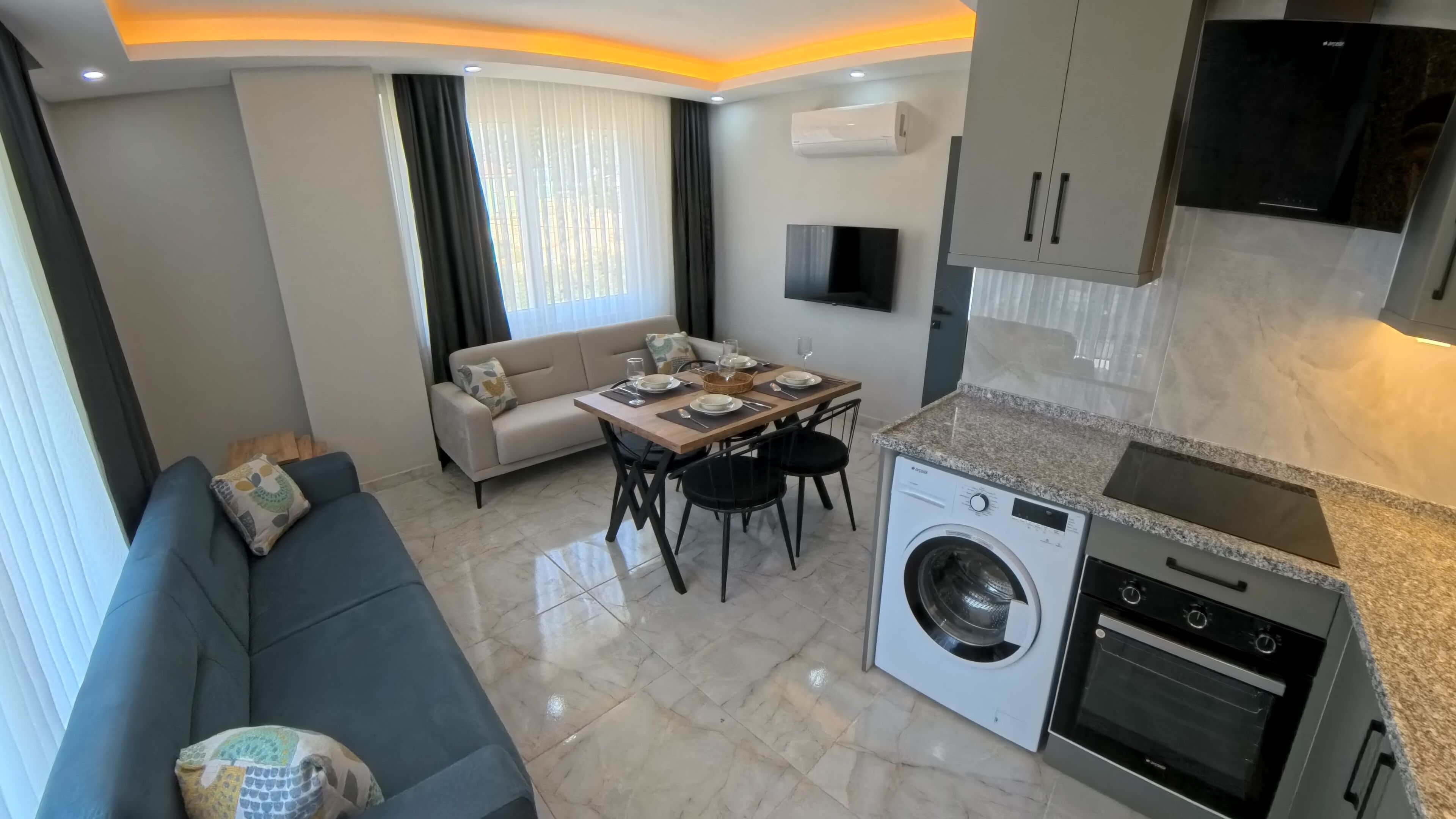 Eine moderne Küche mit integriertem Waschmaschine und Backofen, ein Essbereich mit Tisch und Stühlen sowie ein Wohnbereich mit Sofa und Fernseher.