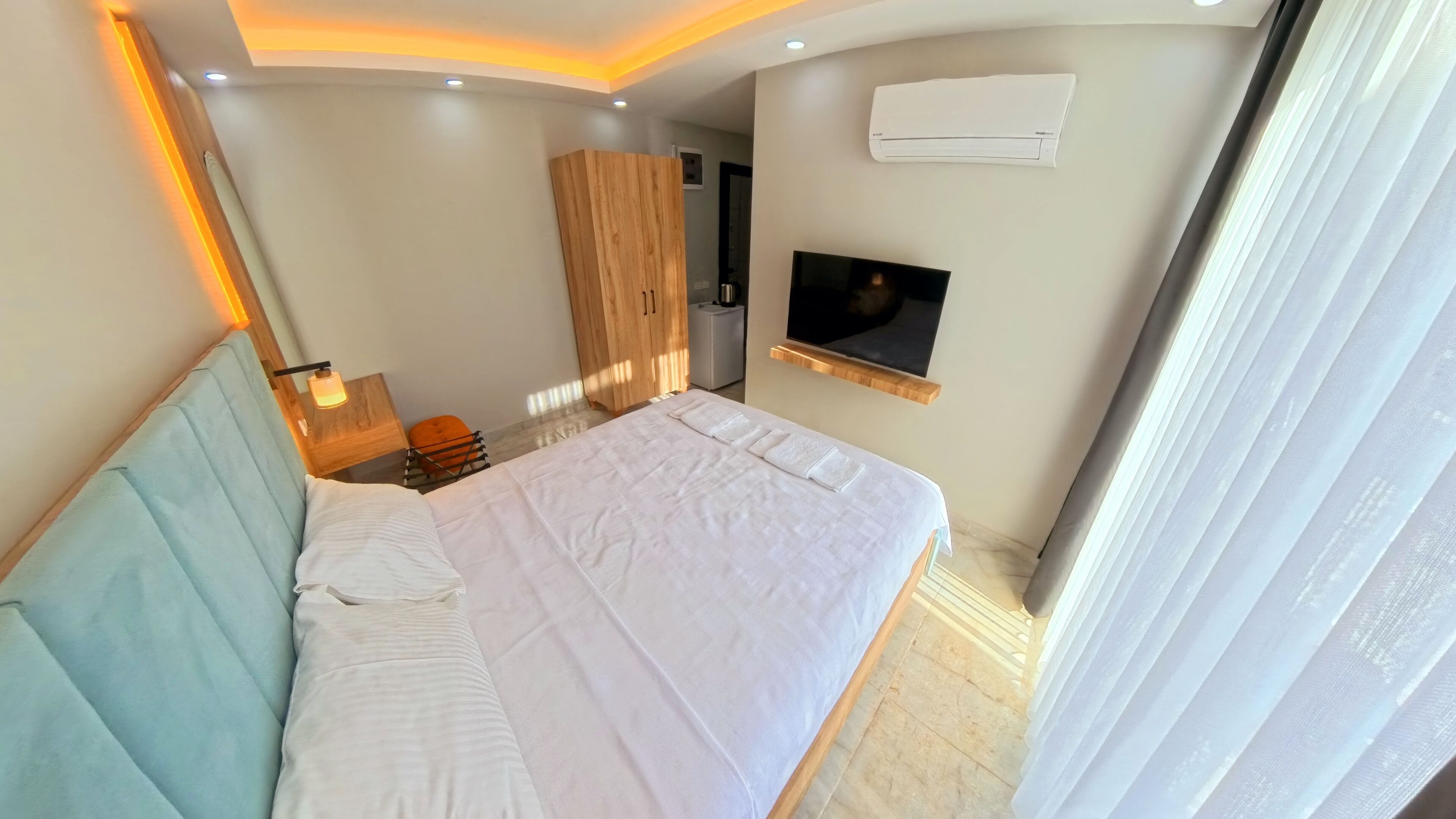 Une chambre d’hôtel moderne, avec un lit bien fait, un téléviseur fixé au mur et une grande fenêtre avec des rideaux.