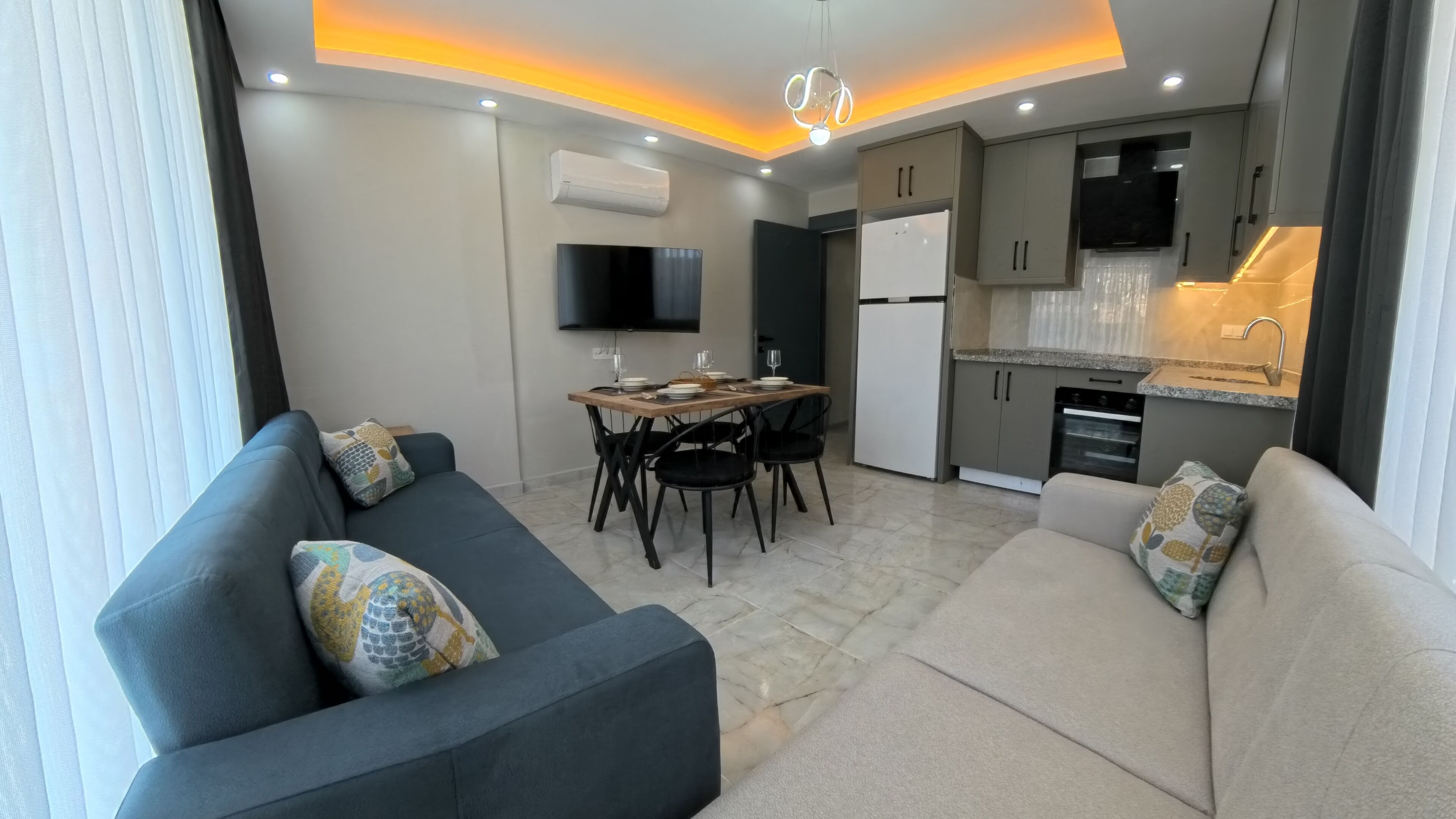 A modern living room with a sofa, a dining table set for four, and a kitchen area with a refrigerator and oven.