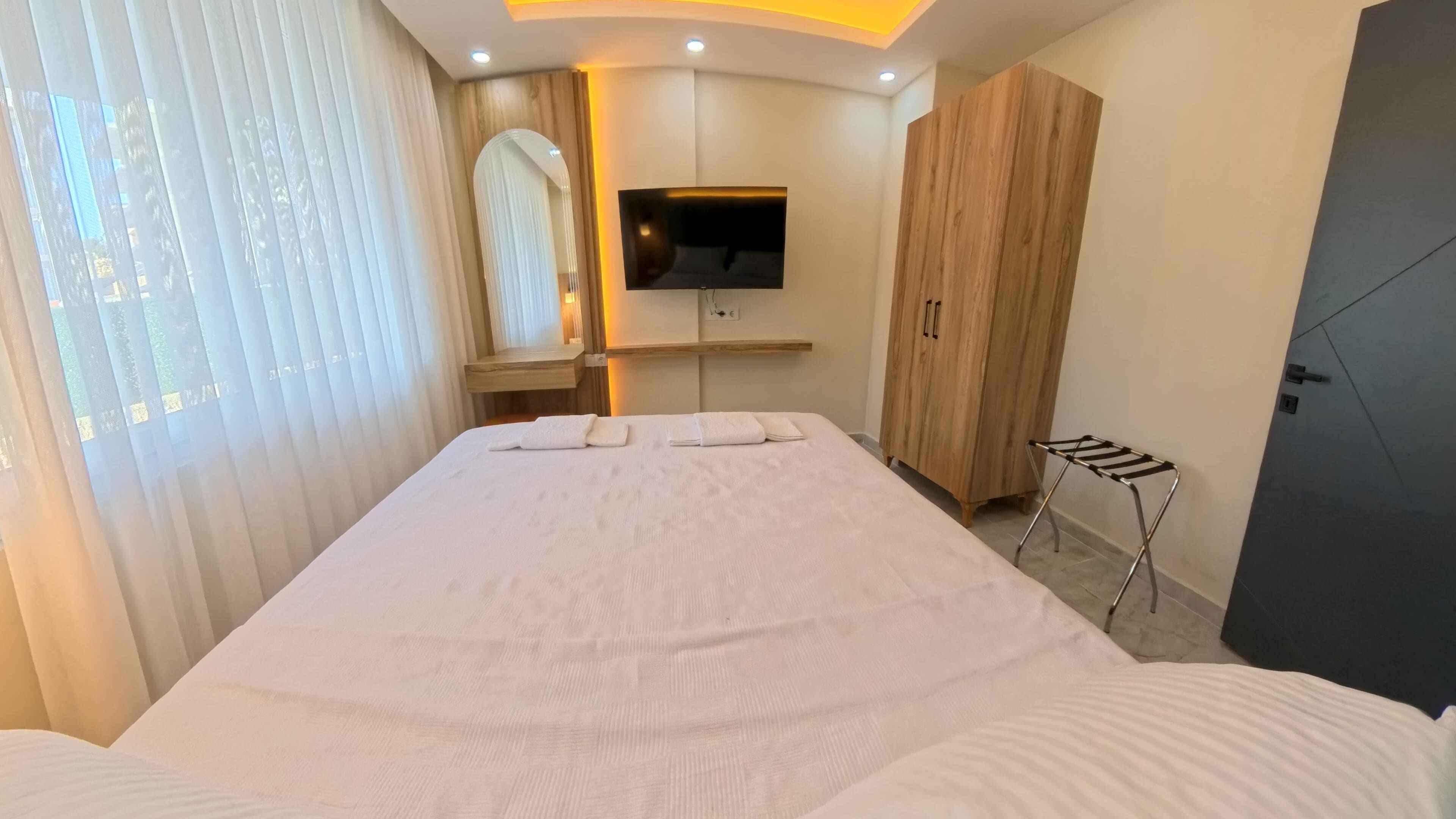 A modern hotel room with a large bed, a TV mounted on the wall, a wooden wardrobe, and a small seating area with a towel.
