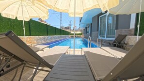 Area piscina con lettini e ombrelloni, una piscina blu cristallina e sullo sfondo edifici.